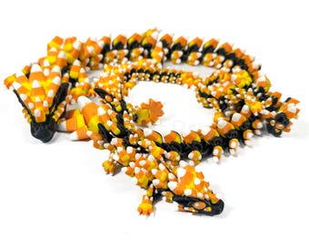May include: A 3D-printed dragon sculpture in the shape of candy corn. The dragon is primarily orange, yellow, white, and black. The body is segmented, and the head and tail are detailed. A unique Halloween decoration.