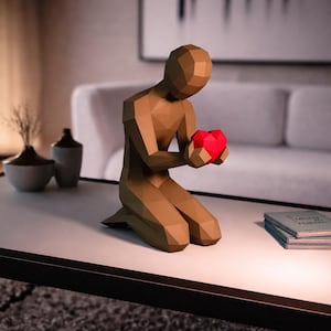 Puede incluir: Una escultura geométrica marrón de una figura arrodillada sosteniendo un corazón rojo. La figura está sobre una mesa blanca, con un fondo borroso con un sofá blanco y jarrones decorativos. La escultura es una obra de arte moderna.