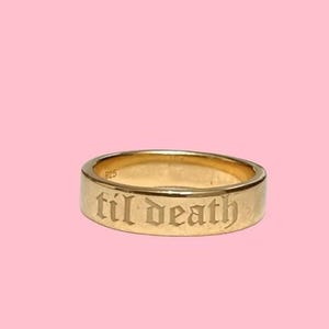 Könnte beinhalten: Ein goldfarbener Ring mit der Inschrift "til death" in einer altenglischen Schriftart. Der Ring ist vor einem rosa Hintergrund platziert. Der Ring ist im Bandstil mit flacher Oberfläche.