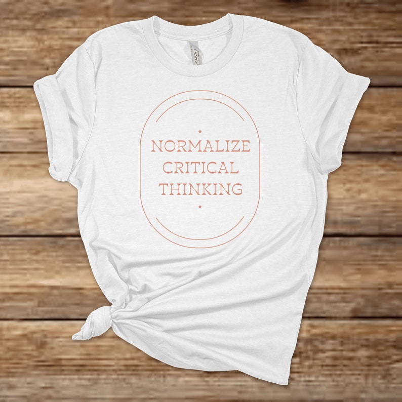 May include: White t-shirt with a brown circle graphic that says "Normalize Critical Thinking".
