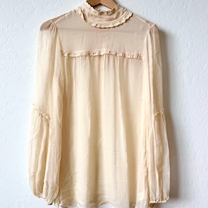 K&ouml;nnte beinhalten: Eine cremefarbene Bluse mit hohem, ger&uuml;schtem Kragen und langen, weiten &Auml;rmeln. Der transparente Stoff h&auml;ngt an einem Holzb&uuml;gel vor einer wei&szlig;en Wand. Die Bluse hat eine zarte, Vintage-&Auml;sthetik.