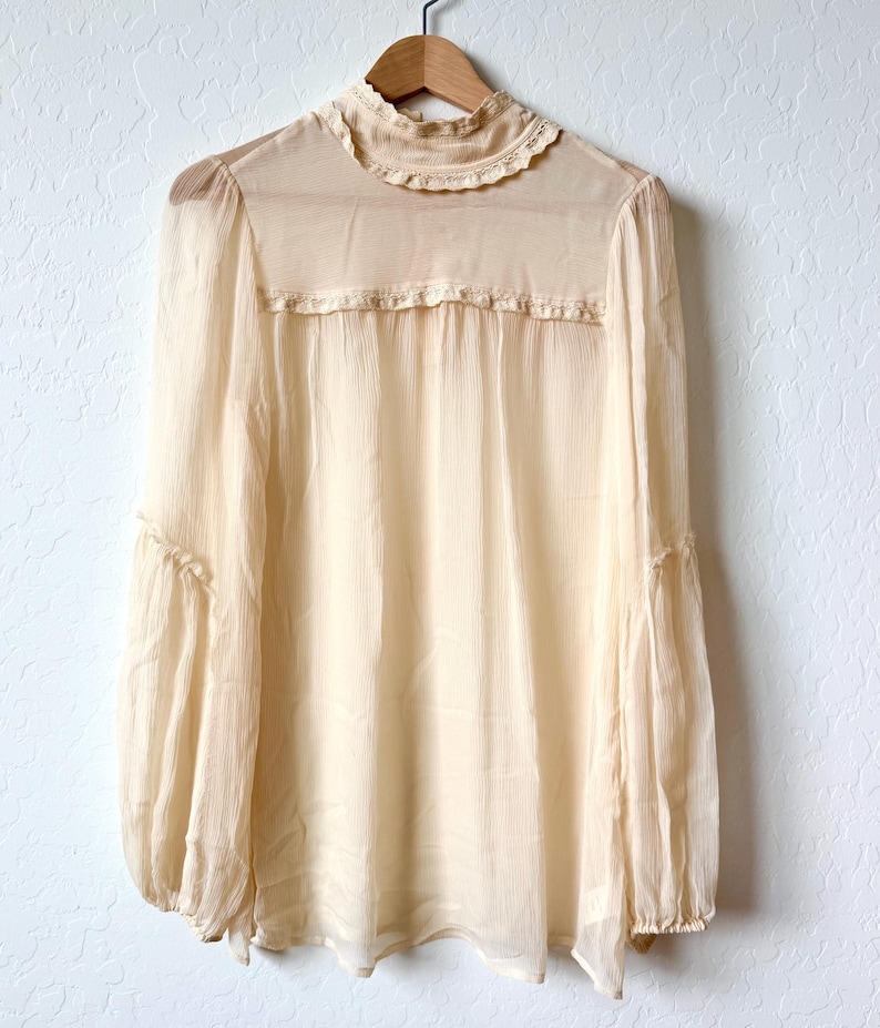 K&ouml;nnte beinhalten: Eine cremefarbene Bluse mit hohem, ger&uuml;schtem Kragen und langen, weiten &Auml;rmeln. Der transparente Stoff h&auml;ngt an einem Holzb&uuml;gel vor einer wei&szlig;en Wand. Die Bluse hat eine zarte, Vintage-&Auml;sthetik.