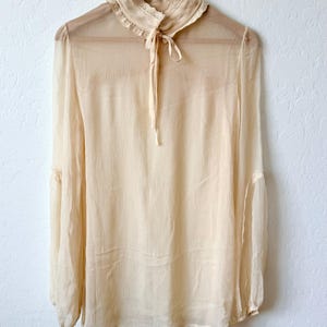 K&ouml;nnte beinhalten: Eine transparente, cremefarbene Bluse mit langen &Auml;rmeln und einem hohen, ger&uuml;schten Kragen. Die Bluse hat eine zarte Schleife am Ausschnitt und h&auml;ngt an einem Holzb&uuml;gel. Der Stoff wirkt leicht und flie&szlig;end, was auf einen Vintage- oder Boh&egrave;me-Stil hindeutet.