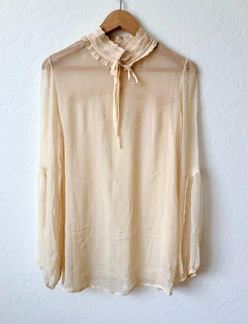 K&ouml;nnte beinhalten: Eine transparente, cremefarbene Bluse mit langen &Auml;rmeln und einem hohen, ger&uuml;schten Kragen. Die Bluse hat eine zarte Schleife am Ausschnitt und h&auml;ngt an einem Holzb&uuml;gel. Der Stoff wirkt leicht und flie&szlig;end, was auf einen Vintage- oder Boh&egrave;me-Stil hindeutet.