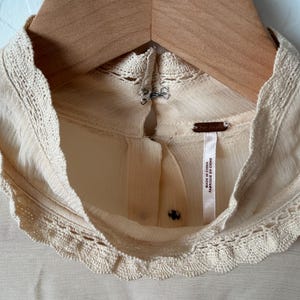 K&ouml;nnte beinhalten: Cremefarbene Bluse mit hohem, rundem Ausschnitt und zarter Spitzenborte. Die Bluse h&auml;ngt an einem Holzkleiderb&uuml;gel. Das Etikett lautet "Made in China / Fabriqu&eacute; en Chine".