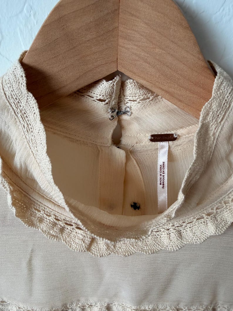 K&ouml;nnte beinhalten: Cremefarbene Bluse mit hohem, rundem Ausschnitt und zarter Spitzenborte. Die Bluse h&auml;ngt an einem Holzkleiderb&uuml;gel. Das Etikett lautet "Made in China / Fabriqu&eacute; en Chine".