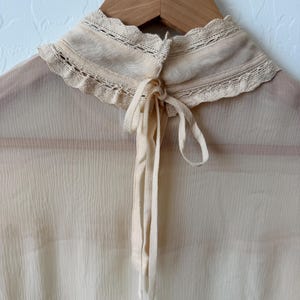 K&ouml;nnte beinhalten: Eine cremefarbene Bluse mit hohem, ger&uuml;schtem Kragen und zarter Spitzenborte. Die Bluse hat einen Bindverschluss am Hals und ist aus einem transparenten, leichten Stoff gefertigt. Das Kleidungsst&uuml;ck h&auml;ngt an einem Holzkleiderb&uuml;gel.