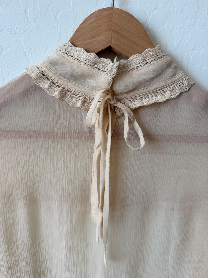 K&ouml;nnte beinhalten: Eine cremefarbene Bluse mit hohem, ger&uuml;schtem Kragen und zarter Spitzenborte. Die Bluse hat einen Bindverschluss am Hals und ist aus einem transparenten, leichten Stoff gefertigt. Das Kleidungsst&uuml;ck h&auml;ngt an einem Holzkleiderb&uuml;gel.
