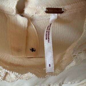 K&ouml;nnte beinhalten: Nahaufnahme einer cremefarbenen Bluse mit Schl&uuml;sselloch-Ausschnitt und zarter Spitzenborte. Das Etikett lautet "Free People" und "Made in China". Die Bluse hat ein Etikett mit der Gr&ouml;&szlig;e 8.