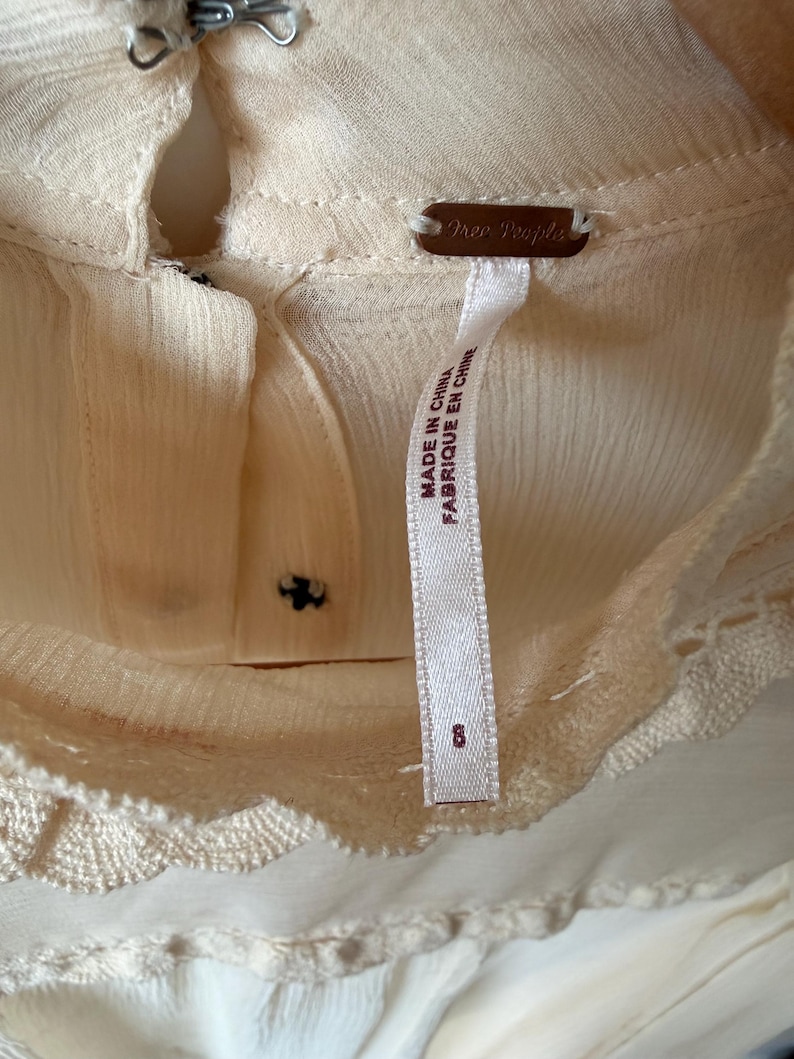 K&ouml;nnte beinhalten: Nahaufnahme einer cremefarbenen Bluse mit Schl&uuml;sselloch-Ausschnitt und zarter Spitzenborte. Das Etikett lautet "Free People" und "Made in China". Die Bluse hat ein Etikett mit der Gr&ouml;&szlig;e 8.