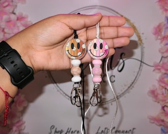 Puede incluir: Dos cordones de cuentas coloridos con un dije de carita sonriente. Un cordón tiene un dije de carita sonriente rosa y blanco con cuentas rosas y blancas. El otro cordón tiene un dije de carita sonriente marrón y blanco con cuentas marrones y blancas.