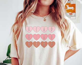 Puede incluir: Una camiseta blanca con la palabra "READ" escrita en corazones rosas y marrones.