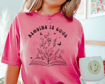 May include: A pink t-shirt with the text "Reading is Cool" in a retro font. The text is surrounded by a floral design with a book open underneath.
