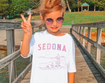 May include: A young person wearing a white t-shirt with the word "SEDONA" in pink and purple lettering. The shirt features a desert landscape design. The person is wearing round sunglasses and giving a peace sign.
