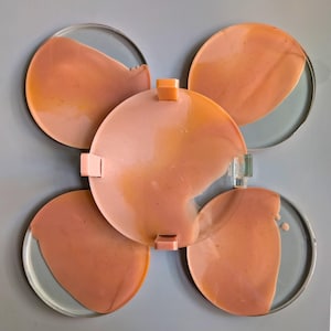 Puede incluir: Obra de arte abstracta que presenta cinco elementos circulares. La pieza central es un círculo más grande con un pasador cuadrado. Cuatro círculos más pequeños lo rodean, cada uno con un borde transparente y un interior de color melocotón semitransparente. Sobre fondo gris.