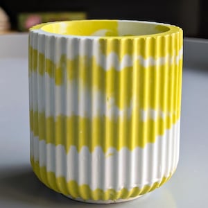 Könnte beinhalten: Ein zylindrischer Pflanztopf mit vertikalen Rillen, bemalt in abwechselnden weißen und gelben Bändern. Der Innenraum ist gelb mit weißen Marmorierungen. Dieser dekorative Behälter eignet sich für kleine Pflanzen oder als Schreibtisch-Organizer.