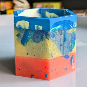Könnte beinhalten: Ein mehrfarbiger sechseckiger Behälter mit einem geschichteten Design. Die Oberseite ist blau, gefolgt von Gelb und dann einer leuchtend orangefarbenen Basis. Die Farben sind gewirbelt und gesprenkelt, was einen einzigartigen, künstlerischen Look erzeugt. Der Behälter dient wahrscheinlich zum Aufbewahren kleiner Gegenstände.