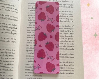 May include: A pink bookmark featuring a repeating pattern of red strawberries and small white hearts. The bookmark is made from a soft, flexible material and has a slightly textured surface. The text "An Offer from a Gentleman" is visible on the page behind the bookmark.