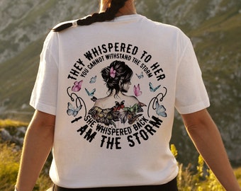 May include: A white t-shirt with a graphic print of a woman with butterflies and flowers. The text on the shirt reads "They whispered to her you cannot withstand the storm. She whispered back I am the storm."