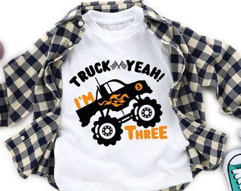 May include: White t-shirt with a black monster truck graphic and the text "I'm Truck Yeah! Three". The shirt is on a checkered shirt and a pair of teal sneakers.