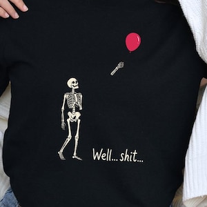 May include: A black t-shirt featuring a graphic of a white skeleton looking up at a detached skeletal hand reaching for a floating red balloon. The text "Well... shit..." is printed in white below the skeleton on the front of the garment.