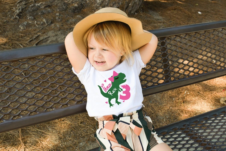 May include: A young child wearing a white t-shirt with a green dinosaur graphic and the text "Three Rex 3" in pink. The child is wearing a brown hat and sitting on a metal bench.