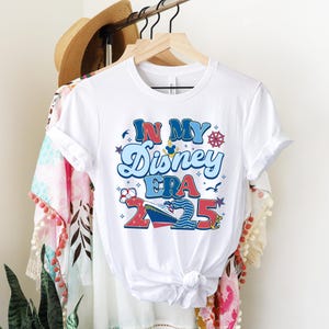 Puede incluir: Camiseta blanca con un gr&aacute;fico colorido que dice "In My Disney Era 2025". El gr&aacute;fico incluye una cabeza de Mickey Mouse, un barco y una br&uacute;jula.