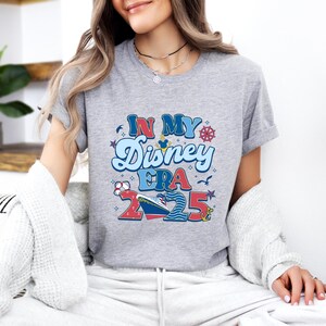 Puede incluir: Camiseta gris con un dise&ntilde;o gr&aacute;fico que dice "In My Disney Era 2025". El dise&ntilde;o presenta un crucero, Mickey Mouse y otros elementos n&aacute;uticos.