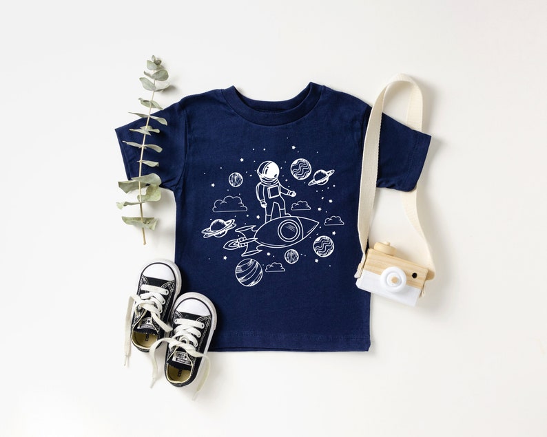 May include: Navy blue toddler t-shirt featuring a white graphic of an astronaut on a rocket ship, with planets and stars. Black and white trainers and a toy camera are also in the image.