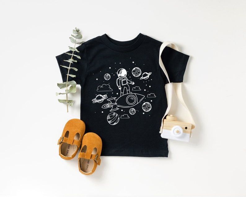 May include: Black toddler t-shirt featuring a white illustration of an astronaut on a rocket, with planets and stars. The image includes brown shoes and a toy camera.