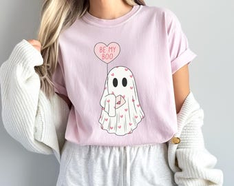 May include: A light pink t-shirt featuring a cartoon ghost holding a letter and a heart-shaped balloon. The balloon reads "BE MY BOO". The ghost is covered in pink hearts.