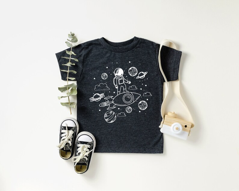 May include: A dark gray t-shirt with a white graphic of an astronaut standing on a rocket in space. The astronaut is wearing a helmet and has a small backpack. There are stars and planets in the background. The t-shirt is on a white surface with a pair of black shoes and a wooden toy camera.