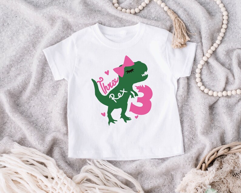 May include: White t-shirt with a green dinosaur wearing a pink bow and the text "Three Rex" and a pink number "3".
