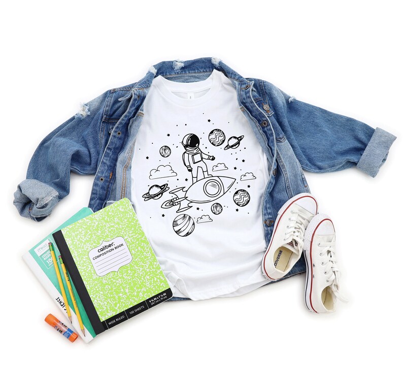 May include: White t-shirt featuring a black and white astronaut and rocket ship design, with planets and clouds. A light blue denim jacket, white trainers, and school supplies are arranged around the shirt.