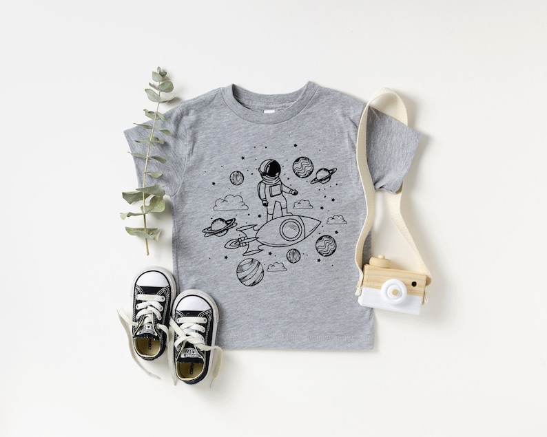 May include: A grey t-shirt with a black and white illustration of an astronaut on a rocket ship, surrounded by planets and stars. The shirt is styled with black and white trainers and a toy camera.
