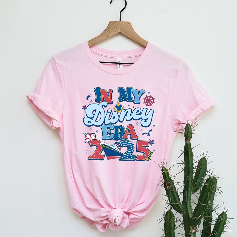 Puede incluir: Camiseta rosa con un dise&ntilde;o gr&aacute;fico que dice "In My Disney Era 2025" con un barco de dibujos animados, una cabeza de Mickey Mouse y otros gr&aacute;ficos de tem&aacute;tica n&aacute;utica.