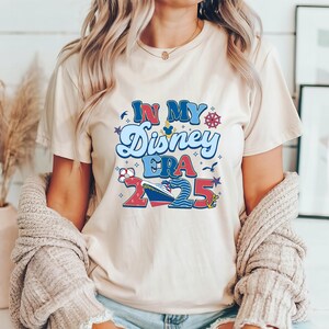 Puede incluir: Una camiseta beige con un dise&ntilde;o gr&aacute;fico que dice "In My Disney Era 2025". El gr&aacute;fico incluye un crucero, una cabeza de Mickey Mouse y elementos n&aacute;uticos como un tim&oacute;n y un ancla.