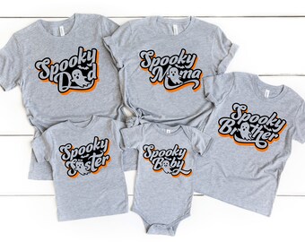 May include: A collection of gray t-shirts and a onesie with the words "Spooky Dad", "Spooky Mama", "Spooky Brother", "Spooky Sister", and "Spooky Baby" in a retro font. Each design includes a ghost and orange accents, perfect for Halloween.