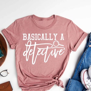 May include: A heather mauve t-shirt with the text "Basically a detective" in white lettering. The shirt is folded and laid flat on a white surface with a pair of glasses, a wooden cutting board, a small potted plant, a vintage camera, a brown wallet, and a pair of blue jeans with white sneakers.