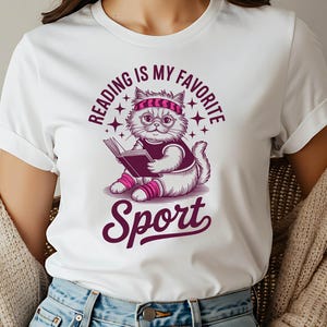 A woman wearing a white t-shirt with a graphic design of a cat reading a book and the text "Reading is my favorite Sport" printed on it.