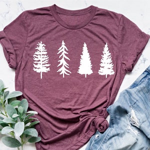 May include: A heather maroon t-shirt with a white graphic of four stylized pine trees. The shirt is laid flat on a white marble surface with a sprig of greenery and a pair of light blue denim jeans with distressed details.