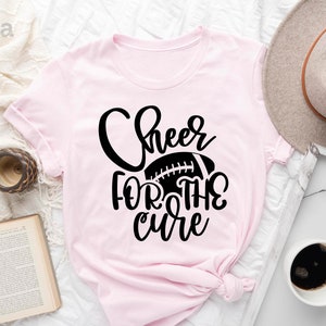 May include: Pink t-shirt with black text that reads "Cheer for the cure" and a black football graphic.
