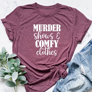 May include: A maroon t-shirt with the text "MURDER shows & COMFY clothes" in white. The shirt is styled with distressed blue jeans. A sprig of green leaves is placed near the shirt and jeans.