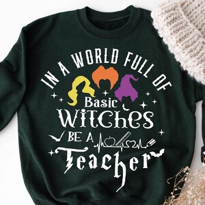 May include: Forest green sweatshirt with the text "IN A WORLD FULL OF BASIC WITCHES BE A TEACHER" in white. The design includes witch silhouettes and other graphics. A cream-colored knit hat and dried flowers are also in the image.