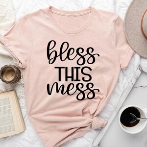 Puede incluir: Una camiseta rosa con el texto "bless this mess" en letras negras.