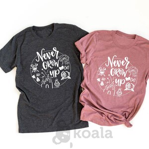May include: Two t-shirts with the text "Never Grow Up" in white lettering on a circle with Disney-themed icons. The shirt on the left is dark heather grey and the shirt on the right is heather mauve.