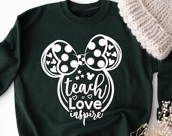 Op de afbeelding: Bosse groen sweatshirt met een witte muizenkop-afbeelding met een strik en de woorden "teach love inspire". Een crèmekleurige gebreide muts en droogbloemen liggen naast het sweatshirt.