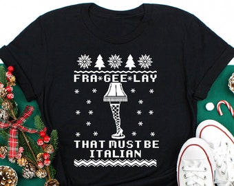 May include: Black t-shirt with a white graphic design. The design includes the text "FRA*GEE*LAY THAT MUST BE ITALIAN" with snowflakes, Christmas trees, and a leg lamp illustration. The shirt is displayed with holiday decorations.