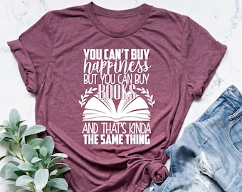 May include: A heather maroon t-shirt with the text "You can't buy happiness but you can buy books and that's kinda the same thing" in white text. The shirt is folded and laid on a white surface with a pair of blue jeans and a green plant.
