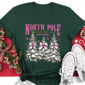 May include: A dark green t-shirt with the text "NORTH POLE CHRISTMAS TREE FARM ALWAYS FRESH" in pink. The shirt features three illustrated Christmas trees with pink bows. White sneakers and holiday decorations are also visible.